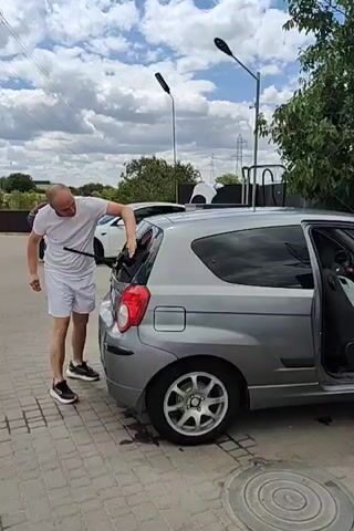 A bald man wearing a white t-shirt and white shorts is standing next to a silver hatchback car. He is holding a long black object, possibly a cleaning tool, and appears to be wiping the rear windshield of the car. The car has its passenger door open, and there are bags visible inside. The man is wearing black sneakers. The scene is outdoors, with a paved surface and trees in the background. The sky is cloudy.