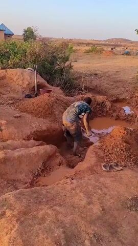 A man is digging in a pit of red clay and water. He is wearing jean shorts, a t-shirt, and is barefoot. He is using a shovel to move the clay and water. The pit is surrounded by piles of red clay. In the background, there is a dry, hilly landscape with some trees and a few distant structures. There are a few tools lying nearby.