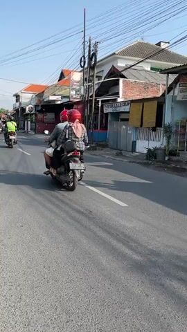 The video shows a street scene with two people riding motorcycles. The motorcycle on the left is ridden by a person wearing a red helmet and a light-colored jacket. The motorcycle on the right is ridden by a person wearing a pink helmet and a light-colored shirt. Both motorcycles are moving away from the camera. The street is paved with asphalt and has white lane markings. On the sides of the street are buildings, some of which appear to be shops. There are trees and power lines visible. The sky is clear and the sun is shining, casting shadows on the road.
