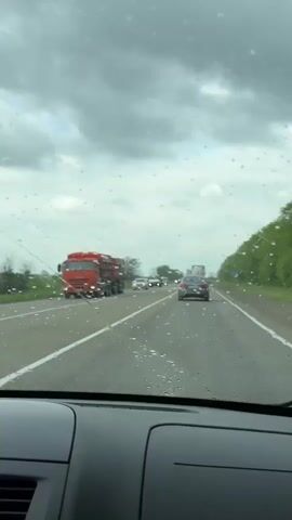 Here is a forensic description of the video:

The video is shot from inside a vehicle, likely a car, looking through the windshield as it travels down a highway. It is raining, and there are visible raindrops on the windshield. The windshield wipers are activated and briefly sweep across the glass. Several other vehicles are on the road, including cars, vans, and a large red truck on the left side of the road. The sky is overcast with grey clouds. Trees line the right side of the road. Road markings, including solid white lines, are visible. The vehicle's dashboard is in the foreground, showing details such as air vents and airbag indicators. Overall, the video captures a scene of driving on a wet highway with moderate traffic under cloudy weather conditions.