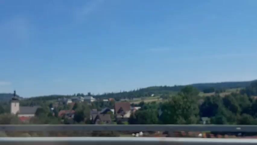The video shows a landscape with a town in the distance. The sky is clear and blue. In the foreground, there is a railing, possibly from a bridge or balcony. Beyond the railing, there are trees and hills. In the town, a church with a steeple is visible, along with several houses. The scene appears to be a sunny day.