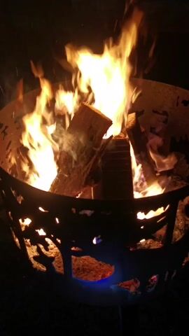 A close-up shot shows a roaring campfire contained within a metal fire pit. The fire pit has a decorative pattern with tree and animal silhouettes cut into its sides. Flames leap high into the air, casting a warm glow on the logs within. The scene is dark, suggesting it is nighttime, and sparks can be seen rising from the fire.