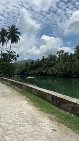 A motorcycle with two people is driving down a dusty road. The road is next to a canal with lush green trees on the opposite bank. There are also coconut trees on the left side of the road. The sky is blue with fluffy white clouds.