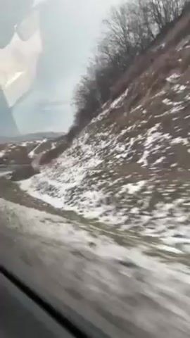 The video shows a snowy landscape from the perspective of someone inside a moving car. The road is partially covered in snow and the adjacent hillside is also blanketed with snow. Bare trees are visible atop the hill. The camera is positioned inside the vehicle, offering a view through the car window. The movement of the vehicle implies travel, and the snowy environment suggests a winter setting. The overall tone is bleak and cold due to the sparse, snow-covered scenery.