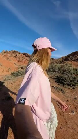 A woman with long blonde hair, wearing a pink baseball cap and a pink oversized t-shirt with a black and white panda logo on the left sleeve, records a selfie video. She also wears sunglasses hanging on her shirt collar and carries a white crocheted bag with fringe over her shoulder. She walks along a dirt path in an arid, mountainous landscape characterized by reddish-brown earth and sparse green bushes. The sky is bright blue with a few wispy clouds. Initially, she looks to her right, then turns her head and smiles at the camera. The camera then pans forward, showing the path ahead, which winds down between the reddish hills. Another person in dark clothing is visible walking further down the path. The scene is bright and sunny, suggesting a clear day. The location appears to be a natural, rugged area, consistent with the provided context of Tosor, Kyrgyzstan.