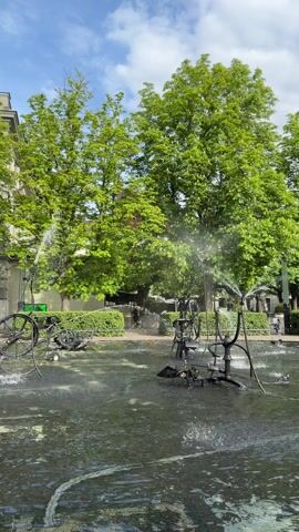 The video shows a fountain containing abstract sculptures constructed from bicycle parts. Water jets spray upwards from various points on the sculptures, creating an active water display. The fountain is set against a backdrop of green trees, a well-manicured hedge, and buildings. People can be seen walking in the background, moving along a paved area beyond the fountain. The weather appears sunny with a mix of blue sky and white clouds visible. The architectural style of the buildings suggests a European setting.