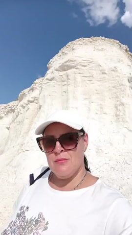 A woman wearing sunglasses and a white baseball cap is taking a selfie. She is standing in front of a large white cliff face, with a blue sky and white clouds in the background. She is wearing a white t-shirt with a floral pattern. The camera is held at chest level, and the woman is looking directly at the camera. The video is shot in landscape mode.