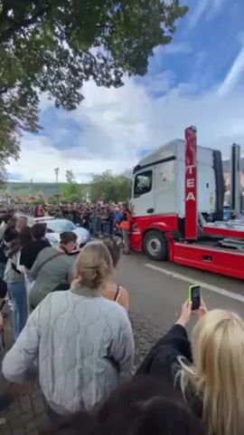 A crowd of people line the street, their cell phones up, recording a white and pale blue Bugatti being driven off of a red and white flatbed truck. The truck is angled towards the left side of the screen. Some of the people are smiling as they watch the Bugatti drive forward. The buildings behind the crowd are multiple stories high and made of stone with brown roofing. The sky is partly cloudy and visible through the leaves of some of the trees on the left side of the screen.