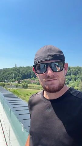 This is a selfie taken by a man with short brown hair and a beard, wearing a black Under Armour baseball cap and black sunglasses. He is also wearing a black t-shirt with a small logo on the left chest that appears to be "4F". He is smiling faintly at the camera.

The setting appears to be outdoors, on a rooftop or elevated platform with a modern glass railing. Behind him, there is a lush green landscape with rolling hills covered in trees. A river or body of water can be seen in the middle ground, with some buildings and boats along its banks. In the distance, on top of a hill, a white tower or building is visible. The sky is clear and blue, suggesting a sunny day.

The time of day is likely daytime, given the bright sunlight and clear sky. The overall mood conveyed by the man's expression and the bright weather is positive and relaxed, possibly indicating a vacation or leisure activity. The location appears to be Hluboká nad Vltavou, Czechia, as suggested by the background landscape and structures. The visible text includes the brand names "UNDER ARMOUR" on the sunglasses and "4F" on the t-shirt.