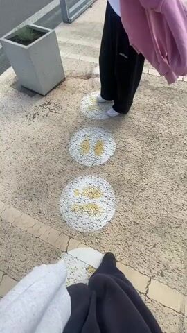 The video opens with a low-angle shot of two people walking on a patterned walkway. The walkway has circular markings with yellow footprints painted on them, indicating a path. The first person, from whose perspective the video seems to be filmed, is wearing black pants and a grey hoodie. Their foot is placed on one of the circular markings. The second person, ahead of the first, is wearing black pants, a white t-shirt, and a pink jacket draped over their shoulder. They are also walking on the marked path. In the background, there is a large blue building with "IKEA" written in large yellow letters. Cars are visible in a parking lot in front of the building, and the sky is partly cloudy. The camera pans slightly to show more of the environment, including a modern metal structure framing the walkway and planters with greenery. The video concludes with a shot of the two people continuing to walk along the path towards the IKEA store.
