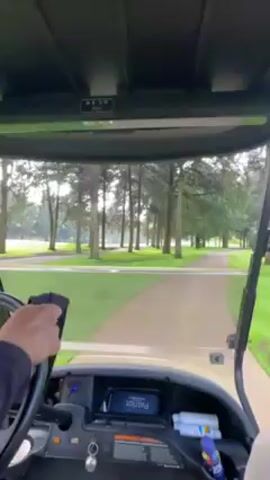 The video shows a first-person perspective from inside a golf cart driving down a paved path on a golf course. The path is lined with trees and leads towards a grassy area. A lake or water hazard can be seen in the distance. The dashboard of the golf cart is visible, with a screen displaying the word "Patriot." A hand holding a black towel is briefly visible near the steering wheel.