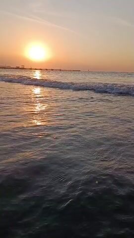 The video shows a close-up view of ocean waves crashing on a beach at sunset. The sun is low in the sky, casting a warm, golden glow on the water. The waves are small and gentle, with white foam crests. In the distance, there is a pier or boardwalk with a few structures on it. The sky is clear and a pale orange color. The camera appears to be stationary, capturing the movement of the water and the light.