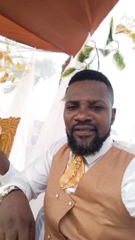 The video captures a man recording with his mobile phone, who is likely at a wedding. He is wearing a white shirt with a gold vest and a gold bow-tie with a gold embellishment. He pans the camera to reveal the bride and groom sitting on a gold ornate couch. The groom is wearing the same vest and bow-tie as the recorder and is using a purple fan. The bride is wearing a white dress. The scene is decorated with white fabric, green vines, and gold flowers. The recorder looks happy.