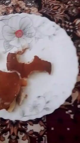 The video shows a close-up shot of a white plate with a floral design on it. The plate contains a partially eaten dessert, possibly pancakes or crepes, with some white powder, likely powdered sugar, sprinkled on top. There are also a few crumbs and a small piece of what appears to be butter on the plate. The plate is placed on a patterned surface, possibly a tablecloth or a bedspread, with dark red and brown tones. The lighting is dim, and the camera is slightly shaky.