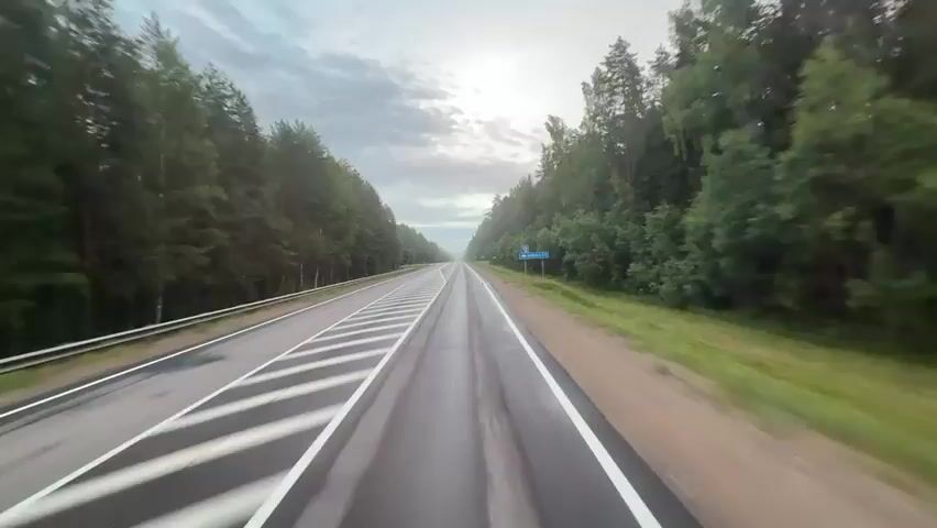 A dashcam view of a two-lane highway flanked by dense green forests. The road surface is wet and marked with white lines, indicating multiple lanes and a dashed center line. The sky above is overcast with patches of light breaking through, creating a slightly hazy atmosphere. A blue road sign is visible on the right side of the road. The camera appears to be mounted on a vehicle moving forward, showing a smooth, straight stretch of road with trees on both sides receding into the distance.