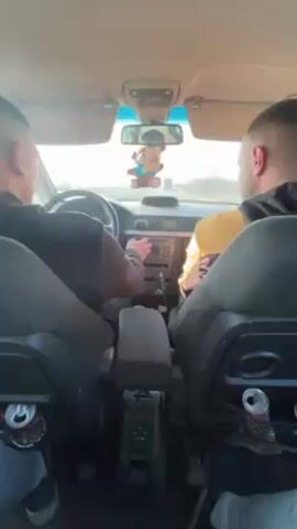 Two men are riding in the front seats of a car driving on a highway in bright sunlight. The driver is in black and the passenger is wearing a yellow jacket. There's a teddy bear hanging from the rearview mirror. Empty beverage cans are visible in the back of the seats. The passenger briefly puts his hand up to block the sun.