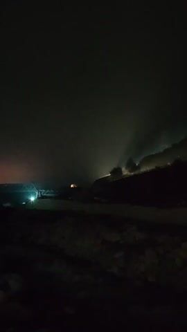 The video shows a nighttime scene with a bridge in the distance illuminated by bright lights. The bridge appears to span a body of water. There is a hill or mountain to the right of the frame with lights visible on its slope, suggesting a settlement or activity. Some of the lights on the hill are diffuse, possibly indicating fog or smoke. The foreground is dark and appears to be uneven terrain, possibly with rocks or debris. The overall atmosphere is dark and somewhat mysterious due to the low light conditions and the presence of fog or smoke.