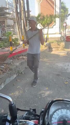 A person wearing a gray shirt, gray pants, and a cap walks from left to right across a dirt path, carrying a broom over their right shoulder. They are walking towards a street with a building on the right side of the frame. The person is the only human visible in the video. The environment appears to be an outdoor urban setting, possibly in a warm climate, given the sunlight. The foreground of the video shows a portion of a motorcycle's dashboard.