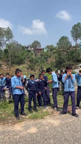 A group of approximately 15 schoolboys in blue uniforms stand together by a road. Some are talking and laughing, while others are eating. The boys are on a grassy area next to a paved road with scattered debris. In the background is a hillside covered with trees and a few houses under a bright, blue sky. One boy walks past the group, wearing a blue jacket and headphones.