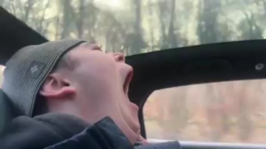 A young man wearing a gray beanie and a dark jacket is sitting in the passenger seat of a moving car.  He yawns widely, then looks toward the camera.  The camera then moves downward toward the legs of someone wearing dark gloves and pants.  A car window and forested scenery can be seen passing by.