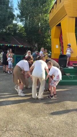 This is a short video clip filmed outdoors in what appears to be an amusement park or recreational area, possibly in Kamchatka, Russia, given the context. The scene is set during daylight, with ample natural light suggesting it's daytime, perhaps late afternoon given the shadows. The weather seems pleasant, as people are dressed in casual summer attire.

The main subjects are a group of people, including adults and children, gathered in a circle on a paved ground. They are actively participating in what looks like a game or a dance, as they are holding hands and bending their bodies in a synchronized movement. One person, a woman in a white shirt and beige pants, is at the center of the group, seemingly leading the activity. Another person, a man in a black shirt, is also part of the circle, leaning in. A young boy in a white t-shirt and colorful shorts is on the right side of the circle, actively engaged.

In the background, there are other people observing the activity. Further back, there's a large, vibrant yellow structure that resembles a playhouse or a themed attraction, possibly a giant wooden horse, hinting at a children's entertainment venue. There are also some trees providing shade and a small building with colorful banners. A speaker is visible on a raised platform next to the yellow structure, suggesting some form of organized entertainment or performance.

A young girl in a patterned outfit and a hat is standing on the platform, looking towards the group. The overall atmosphere appears joyful and energetic, with the participants seemingly having fun. There is no visible text in the clip.