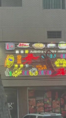 The video captures a storefront displaying a colorful neon sign with Asian food imagery and text. The sign features various seafood and food illustrations, including a large red lobster, a yellow crab, a fish, and dishes of food. The text, partially visible, reads "Auténtica cocina asiática" and "Especialidad en mariscos," indicating authentic Asian cuisine and seafood specialties. Below the neon sign, a glass storefront reveals posters of food items, suggesting a restaurant or food establishment. The building's exterior is brick, with windows visible on the upper floors. A white vehicle is parked in front of the establishment. The lighting suggests it might be daytime or evening with artificial lights on.