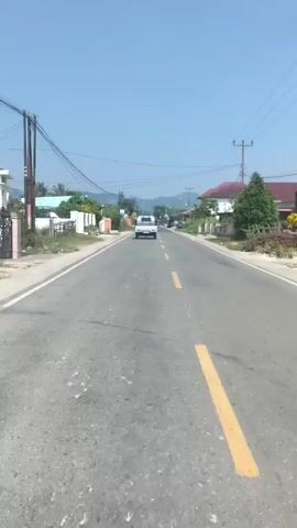The video shows a view from a vehicle traveling on a paved road on a sunny day. The road has a yellow line marking the center. A white car is visible in the distance, traveling in the same direction. On the left side of the road, there is a white fence, some greenery, and buildings. On the right side of the road, there are houses with red roofs and trees. Utility poles with wires are visible along the sides of the road, and mountains are in the background.