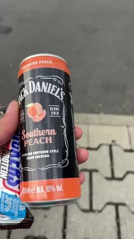 A person's hand holds a can of Jack Daniel's Southern Peach cocktail and a Snickers Protein bar. The can is black with orange accents and features the Jack Daniel's logo, a peach graphic, and the text "SOUTHERN PEACH," "FRESH TASTE," "SERVE COLD," and "SPARKLING SOUTHERN STYLE PEACH COCKTAIL." It also indicates "330 ml e ALK. 10% Vol." The Snickers bar is in its wrapper, which has blue and white text, including "PROTEIN" and "30% PROTEIN." The background shows a gray asphalt surface with a textured, light-colored paved area that appears to be a sidewalk or pathway. The scene is outdoors, and the lighting suggests it might be daytime, possibly overcast given the diffused light. The overall impression is casual, perhaps a person stopping for a snack and a drink.