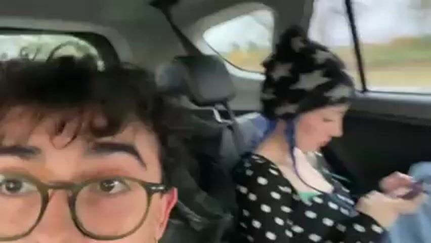 A handheld video shows the interior of a car in motion, with a person in the front passenger seat recording. The person in the front passenger seat has dark, curly hair, is wearing round glasses, and has a silver hoop earring. A person sits in the back seat. They are wearing a black and white polka-dot shirt and a black and grey star beanie with blue braids. The person in the back seat is looking down at a phone. The car is driving past a rural area with a cloudy sky and power lines. Music is playing.