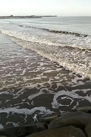 The video shows a calm ocean with gentle waves lapping at the shore. The water is clear and the sand is dark. In the foreground, there are some large rocks, and in the distance, a faint shoreline with some structures is visible. The sky is overcast, and the overall mood is peaceful.