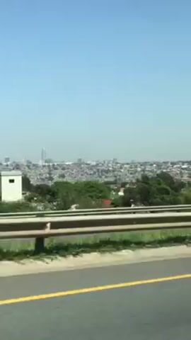 The video, shot from a moving vehicle on a highway, shows a cityscape in the distance under a clear blue sky. The camera passes by a highway sign with four arrows pointing straight ahead, with one arrow branching off to the left. A billboard advertising a Tata Punch car is then visible.  A white pickup truck with a black roll bar and a man standing in the bed drives past. The highway is bordered by a metal guardrail and grassy foliage.