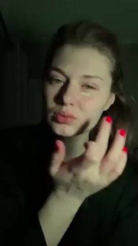 A fair-skinned young woman with blue eyes and long reddish-brown hair is applying a clear product under her left eye. Her nails are painted red. She is wearing a black top or robe. The lighting changes from dim to blue to purple, creating a stylized effect.  The background is dark, with a hint of a door frame visible. The woman's expression is somber, and the focus is on her face and hand as she applies the product.