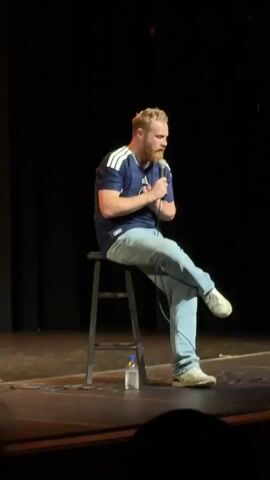 A man with a beard and blonde hair, wearing a dark blue t-shirt with white stripes on the sleeves and light blue jeans, is sitting on a black stool on a stage. He is holding a microphone and talking, with his legs crossed. A clear plastic bottle of water is on the floor next to the stool. The background is dark and appears to be the back wall of a theater or stage. The man is gesturing with his hands as he speaks.
