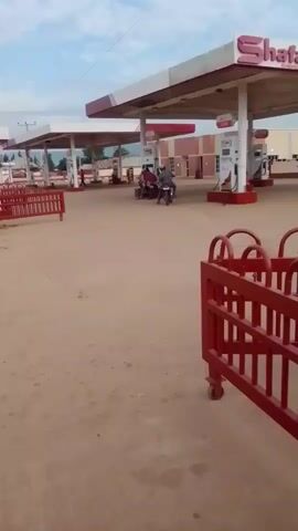 The video shows a gas station with multiple pumps under a canopy. The ground is dirt and there are red metal barriers in the foreground and middle ground. There are a few people and motorcycles at the gas station. In the background, there is a building and some trees. The sky is cloudy.