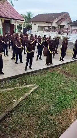 A group of school-aged girls, all wearing hijabs and dark uniforms with yellow accents, are performing synchronized movements outdoors. They appear to be engaged in some form of exercise or dance routine. The background shows a school building with a red roof and some trees. The ground is paved in the area where the girls are standing, with grass and some debris visible in the foreground. The sky is overcast.