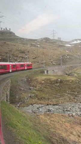 The video shows a red train traveling through a mountainous landscape. It is overcast and rainy, with water droplets visible on the window. The train tracks curve around the side of a mountain, next to a small stone shed. A stream runs below the tracks. Patches of snow are visible on the higher elevations. The view is from inside a train looking out the window.