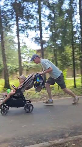 A man is pushing a stroller with a child inside on a paved path in a wooded area. The man is wearing a light-colored t-shirt, dark shorts, and a baseball cap. He appears to be jogging or walking briskly. The stroller is dark-colored and has a bag attached to the handle. The child in the stroller is mostly obscured but appears to be wearing a green shirt. The path is lined with trees and vegetation. Another person, a woman, is walking in the opposite direction on the left side of the path, carrying a phone. The video is shot from a perspective that follows the man and stroller.