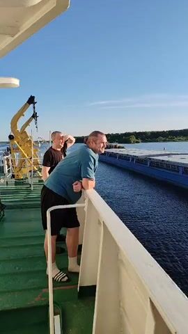 The video captures two individuals on the deck of a vessel, looking out at a passing barge on a sunny day. The person in the foreground, wearing a teal t-shirt and black shorts, leans on a railing, smiling as they observe the barge. To their left, another person in a black t-shirt and shorts shields their eyes, also watching the barge.

The barge, a long, flat-bottomed vessel, is blue with a lighter blue hull and a light-colored deck, suggesting it is transporting goods or materials. In the background, a tree-lined shore is visible, indicating a waterway such as a river or canal. The sky is clear blue with a few wispy clouds, suggesting pleasant weather. The time of day appears to be late afternoon or early evening, given the warm, golden light.

A yellow crane is partially visible on the deck of the viewer's vessel, suggesting it is a working ship. The water is calm and reflects the blue sky. The overall mood seems relaxed and observational, with the individuals enjoying the scenery and the passing of the barge.

No visible text is present on the barge or the surrounding environment that can be discerned from the video. However, the presence of a large barge on a waterway in Russia, specifically near Sud'bitsy, suggests potential industrial or commercial activity related to water transport.