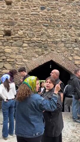 A crowd of people gather around an arched entrance in a tall stone wall. Most stand and chat amongst themselves in groups, while some walk by the entrance. The people are dressed in everyday clothing, and they have different heights, builds, and hairstyles. One woman, dressed in a green headscarf and denim jacket, appears to be speaking to a woman in a black jacket.  The arched entrance has a lightbulb hanging inside. The wall is made of rough stones and mortar. The sky is visible above the wall.