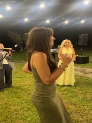 The video captures an outdoor evening event, likely a party or wedding reception, illuminated by string lights overhead. The ground is covered in grass with a stone pathway winding through. In the foreground, a woman in a green strapless dress is visible on the left, her face turned to the right and a smile on her face. Further back, a woman in a pale yellow polka-dot dress is dancing, facing the camera. Behind her, a person is seen operating DJ equipment. To the right of the DJ, a man in a white shirt and black pants is visible. Another woman, wearing a short, sparkly white dress, is actively dancing in the center of the frame, facing the camera and gesturing with her hands. Further in the background, a man in a white shirt and a woman in a pink dress are dancing closely together. The overall atmosphere appears festive and lively.