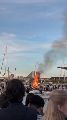 A large bonfire is burning fiercely in an urban waterfront area with a body of water in the foreground and boats docked. Numerous people are gathered on the waterfront and on boats, watching the fire. A building with a smokestack is visible in the background, with smoke from the bonfire rising towards it. The sky is partly cloudy and appears to be dusk or late afternoon. The camera is positioned behind a group of people, suggesting they are part of the crowd observing the event.