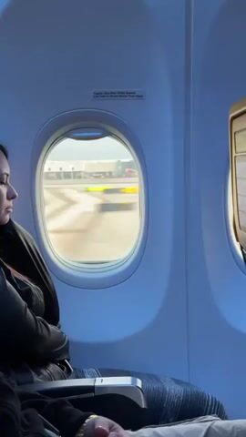 The video shows the interior of an airplane cabin from the perspective of a passenger. Through a small oval window, a runway and airport buildings are visible in the distance. The window also shows what appears to be another airplane on the runway. In the foreground, the back of a passenger's head and shoulder are visible, along with the armrest and part of a seat. To the right of the window, a seatback entertainment screen is visible. The lighting suggests it is daytime. The video captures a moment of an airplane taxiing on a runway.
