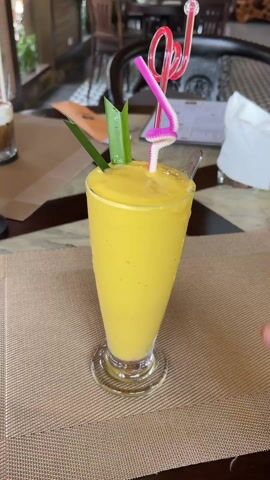 Here is a description of the video:

The video shows a tall glass of a yellow smoothie, likely mango, garnished with a green plant sprig and two straws: one pink and one white. A person is using a spoon to stir and lift some of the smoothie to taste or show its texture. The drink sits on a brown woven placemat on a table in what appears to be a restaurant. In the background, there are other tables and chairs, with a partial view of a dark-colored beverage in another glass. The table has a decorative brown and black pattern. The focus of the shot is clearly on the mango smoothie.
