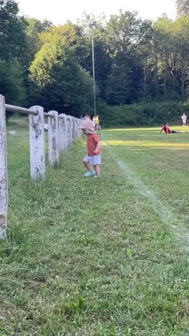 A young child wearing an orange shirt, white shorts, a light-colored hat, and blue shoes walks across a grassy area next to a white-fenced barrier. In the background, a person walks away from the camera, holding a dark object. Further back, several people are gathered, some sitting, near a white line on a green field. The scene is outdoors with trees and foliage in the background.