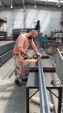 A man with a beard and tattoos on his arms and legs is using an angle grinder to cut a long metal bar. He is wearing a pink t-shirt, khaki shorts, black shoes, and safety glasses. Sparks are flying from the grinder as he cuts the metal. He is standing in a workshop, with stacks of metal bars in the background. There is a table with some rolls of material on it, and a metal railing in the foreground.