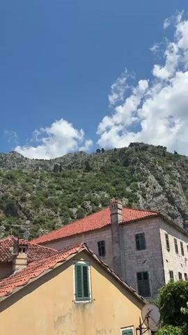 This media file depicts a lively outdoor scene in Kotor, Montenegro, under a bright, sunny sky with scattered white clouds. The foreground features the terracotta tiled roofs of traditional European buildings, one with a prominent yellow facade and green shutters. In the midground, several more multi-story buildings with similar architectural styles, including stone facades and balconies, line a cobblestone plaza.

The plaza is bustling with activity. Numerous people are walking, talking, and enjoying the day. Some are seated at outdoor cafes, shaded by large awnings, suggesting a midday meal or leisurely drinks. The overall atmosphere appears relaxed and pleasant, typical of a tourist destination on a summer day.

In the background, a dramatic mountain range dominates the skyline. The slopes are rugged and covered with dense green vegetation, interspersed with rocky outcrops. A visible fortress or ancient structure can be seen perched high on the mountainside, hinting at the historical significance of the location. The combination of the charming old town architecture and the imposing natural landscape creates a picturesque setting. There is no visible text in the media file.