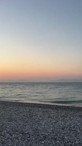This media file captures a serene beach scene at sunset on Rhodes, Greece. The foreground is dominated by a shore of smooth, grey pebbles. Beyond the pebbles, the gentle waves of the Aegean Sea create a mesmerizing pattern of light blue and turquoise hues, reflecting the soft colors of the sky. The water's surface is textured with small ripples, suggesting a light breeze.

In the distance, a faint silhouette of landmasses, likely islands or coastal mountains, can be seen against the horizon. The sky transitions from a pale, clear blue at the top to a warm gradient of soft orange and pinks near the horizon, indicative of the sun setting. The overall atmosphere is peaceful and tranquil, evoking a sense of calm and natural beauty.

No people or specific objects are clearly discernible within the frame, emphasizing the untouched, natural landscape. The time of day is clearly sunset, as evidenced by the vibrant colors in the sky. The location is identified as Rhodes, Greece, which contributes to the visual context of a Mediterranean coastal setting. The emotional tone conveyed is one of serenity, beauty, and perhaps a touch of melancholy associated with the fading light. There is no visible text in the media file.