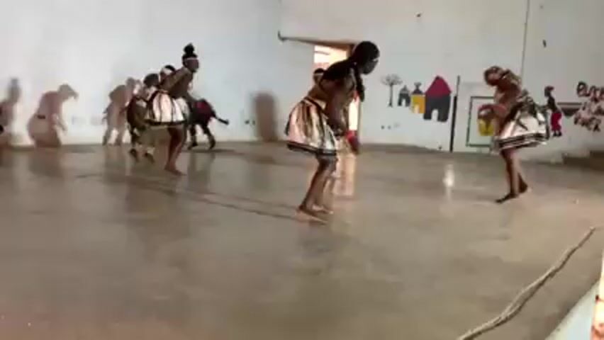 A group of individuals is performing a traditional dance in a large, open room. The dancers are dressed in matching tribal attire, including headbands, beaded necklaces, and short skirts with patterned designs. They are barefoot. The room has white walls, and a door is visible in the background. There's a mural painted on the wall that includes images of huts and stylized figures. The dancers move in sync, performing rhythmic steps and gestures. The camera captures their movements from a low angle, providing a clear view of the dance.