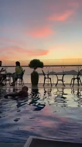 The video shows a man swimming in a pool with a sunset in the background. Two people are sitting at a table by the pool. The pool appears to be on a rooftop terrace with a glass railing. There are additional tables and chairs on the terrace. A potted bush is visible. The sky is multicolored, with shades of pink, orange, and blue. The man in the pool is swimming towards the edge.
