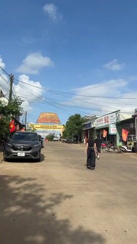 The video captures a sunny daytime street scene in what appears to be a small town or village. The sky is bright blue with scattered white clouds. The street is paved, and power lines crisscross overhead.

In the first shot, a grey SUV is parked on the left side of the road, facing away from the camera. Its license plate "37H 623.11" is visible. Further down the street, an archway spans the road, prominently displaying "CHỢ KON PLÔNG" in large golden letters. Above this, the text "CÔNG TY TNHH MỸ LONG MANG ĐEN" is written, and below, "Hien Giáp Lạc Luỹ Khách". A few people are walking on the right side of the street, including one person in dark clothing and another in a light-colored shirt. Buildings line both sides of the street, with Vietnamese flags hanging from poles.

The camera zooms in on the archway. Other vehicles, including a white SUV and a red truck, can be seen beyond it.

The camera then pans to the left side of the street, showing a storefront with partially visible text "MỘC - SƯ... KIM LIÊN". Two scooters are parked in front of this building, and two red-orange umbrellas shade an outdoor seating area with tables. The building next to it has metal grates covering its entrance.

On the right side of the street, a building has a sign with "QUẢNG 92" and additional Vietnamese text below it. Motorcycles are parked in front of this building.